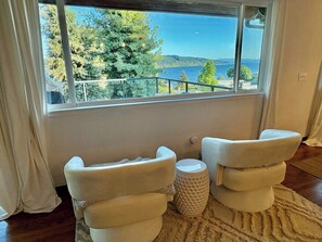 A room with a large window offering a view of trees and a body of water. There are two white chairs and a small round ottoman. - Mt. Rainier & Lake Washington views - cozy 3BR guest suite in Lake Forest Park (Lake Forest Park)