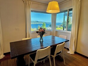Un espace repas comprenant une table, des chaises, un vase avec des fleurs et une vue sur une étendue d’eau, visible à travers de grandes fenêtres.
