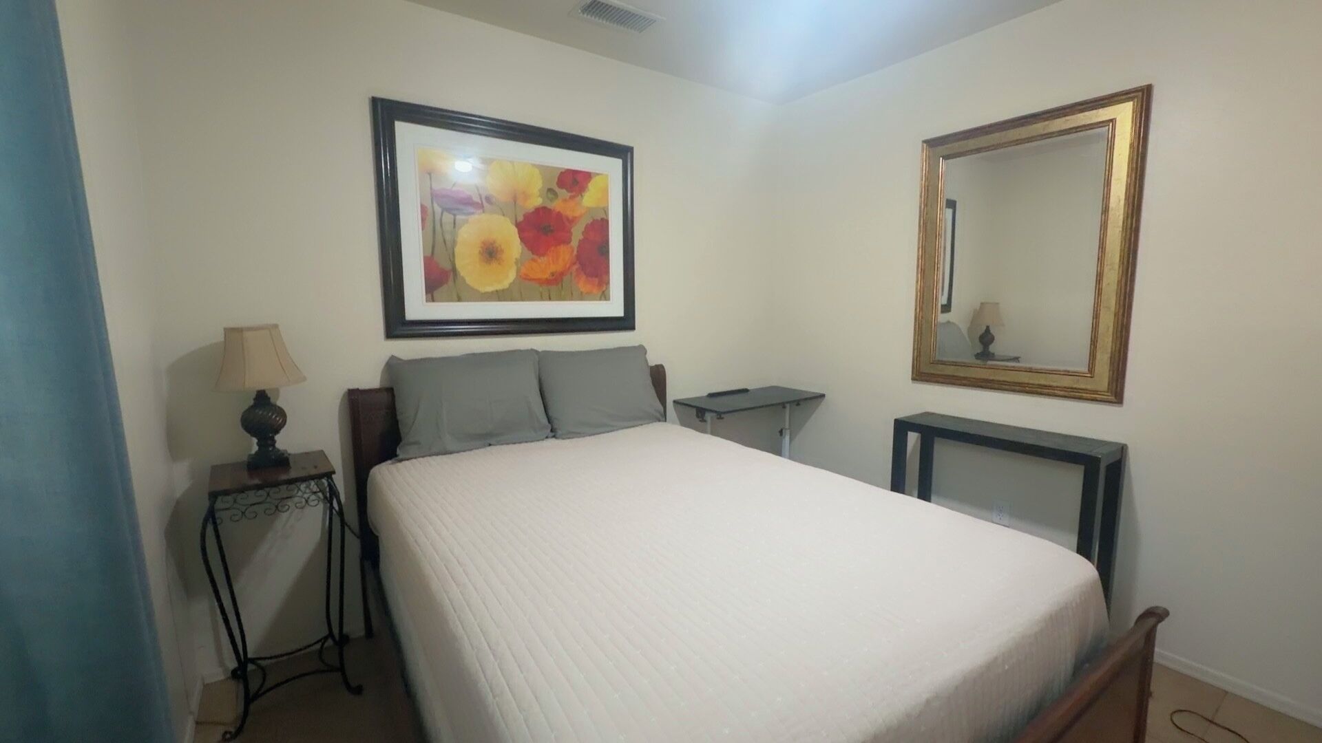 A bedroom with a bed, a bedside table with a lamp, a framed picture, a mirror, and a small table.