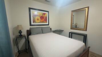 A bedroom with a bed, a bedside table with a lamp, a framed picture, a mirror, and a small table.