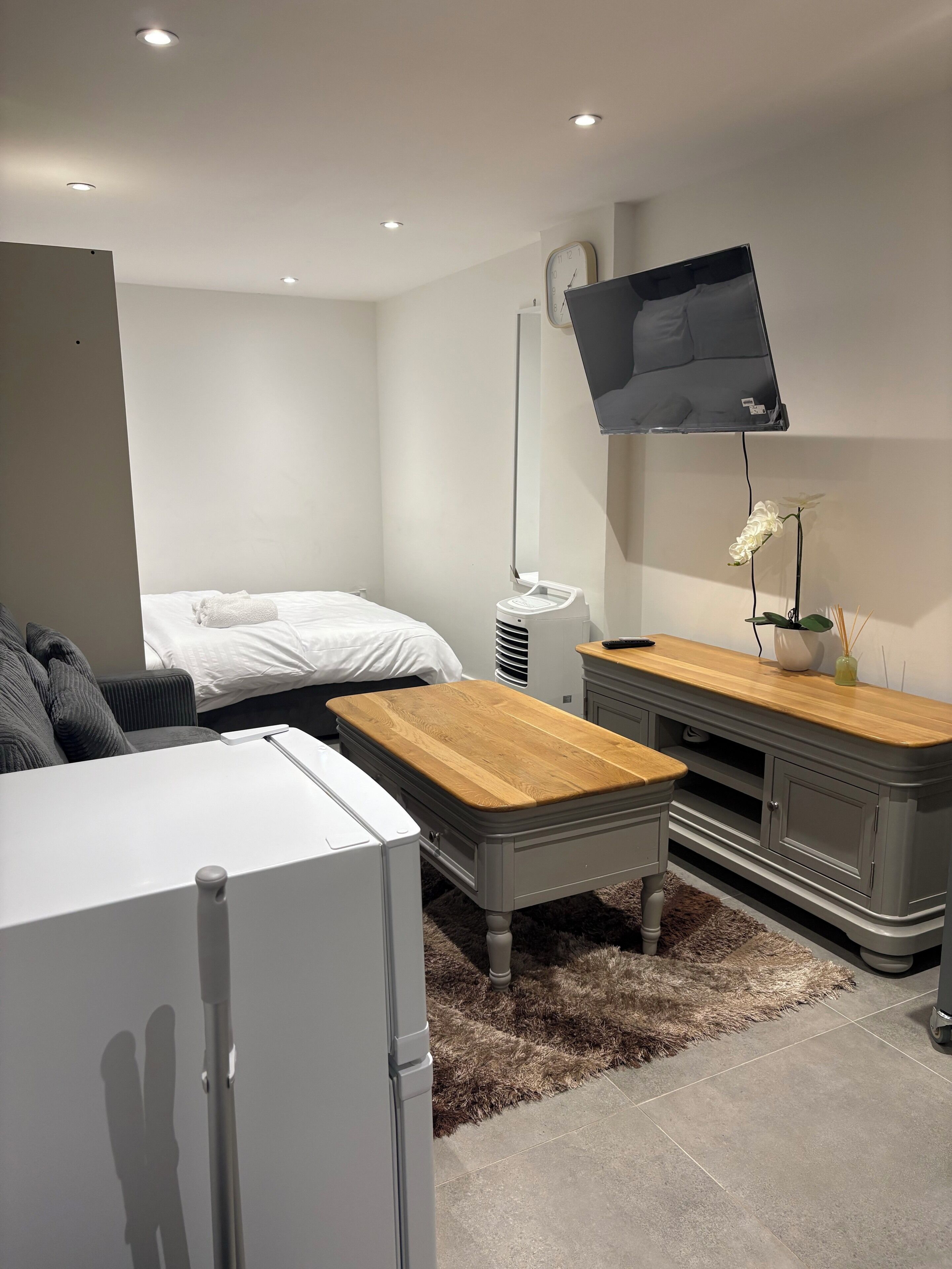 Quarto moderno com cama, frigorífico branco, mesa de madeira, armário cinzento e televisão na parede.