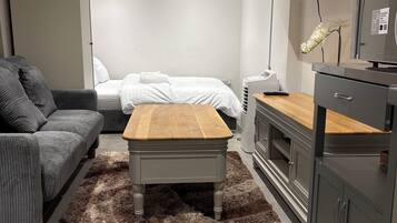 Et moderne soveværelse med en seng, en grå sofa, et træbord, et fjernsyn og et lille skrivebord med en computer.