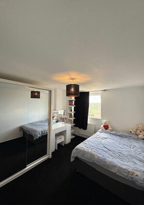 Um quarto com cama, escrivaninha, espelho, luminária pendente e um ursinho de pelúcia.