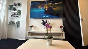 A modern living room with a flat-screen TV mounted on the wall, a white table with a vase of flowers, and shelves with plants. - Apartment Sleeps 2 with Free Parking (Orpington)