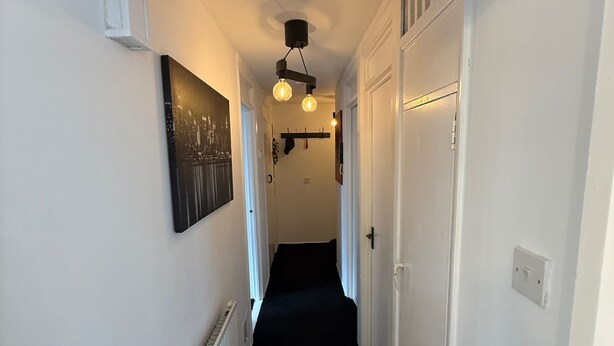 A narrow hallway with a black carpet, white walls, and a cityscape painting. - Apartment Sleeps 2 with Free Parking (Orpington)