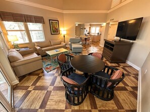 A spacious living room with a checkered hardwood floor, a round dining table with chairs, a sofa, and a television. - Orlando | Island Living Group Stay | Pool + Golf | 3BR Sleeps 12 (Lake Buena Vista)
