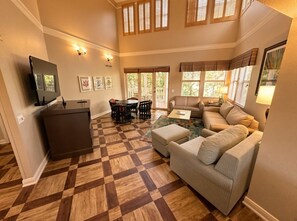 A spacious living room with a checkered floor, a sofa set, a coffee table, a TV, and a dining area with chairs. - Orlando | Island Living Group Stay | Pool + Golf | 3BR Sleeps 12 (Lake Buena Vista)