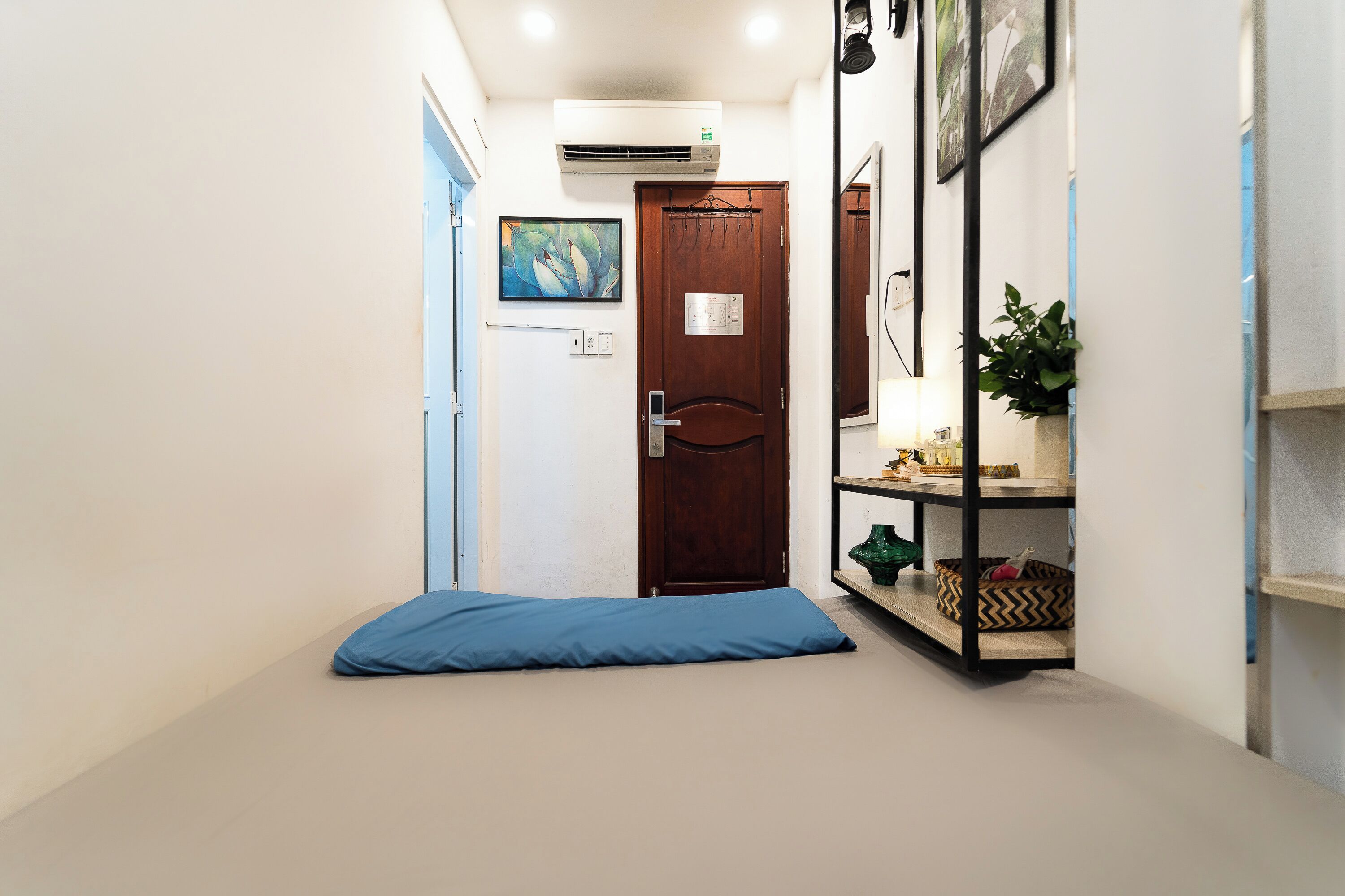 Un pasillo con una puerta de madera, un cojín azul en el suelo, un aire acondicionado de pared y una estantería con objetos decorativos.