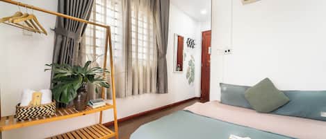 A room with a bed, a wooden shelf with towels and a plant, a window with curtains, and an air conditioning unit.