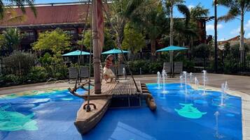 A water feature with a wooden boat and a child playing, surrounded by palm trees and a building with a red roof.