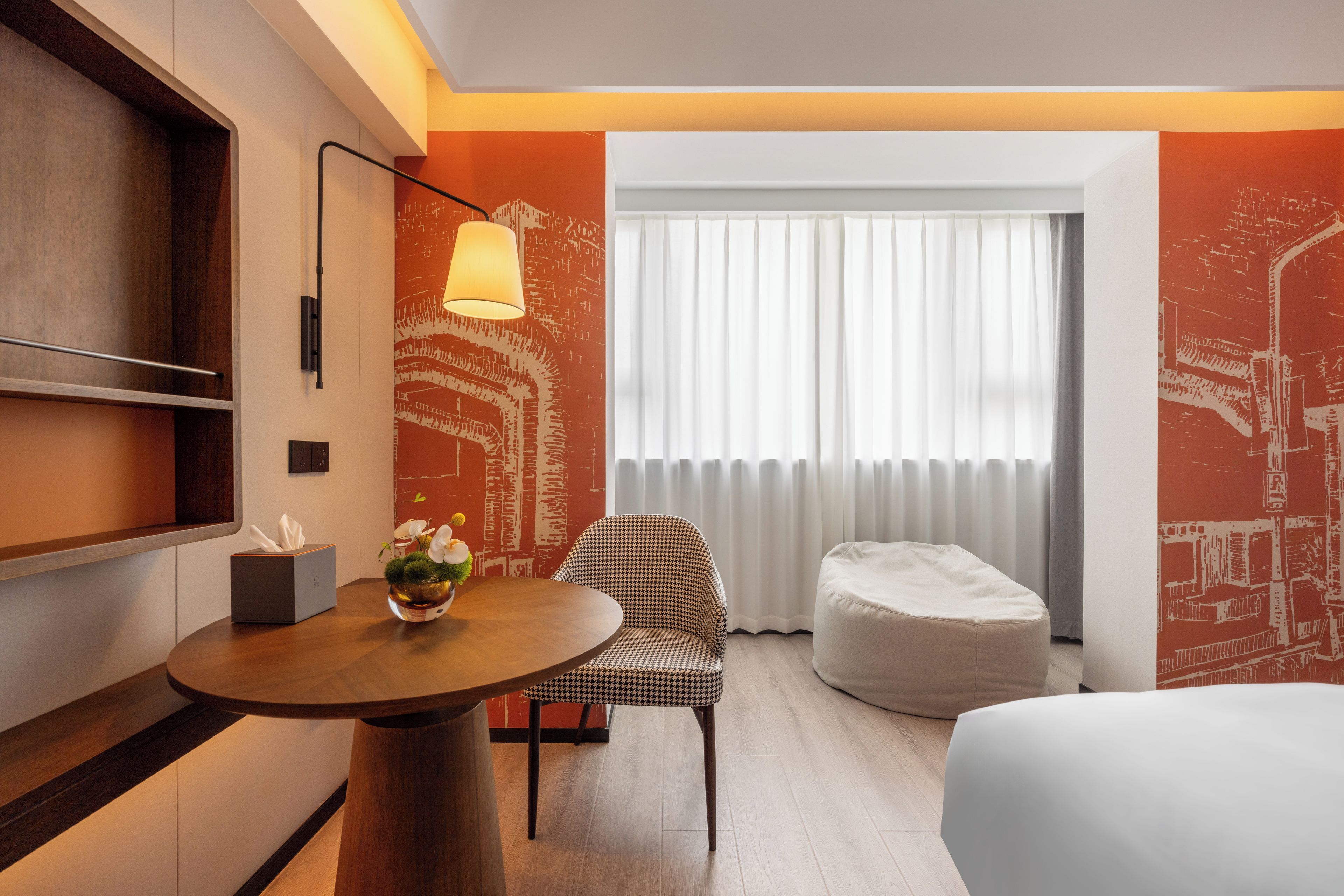 A hotel room with a round wooden table, a houndstooth patterned chair, a white ottoman, and a bed.