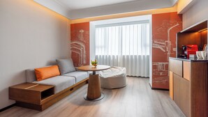 Et moderne hotelværelse med sofa, skammel, rundt bord og et tekøkken.
