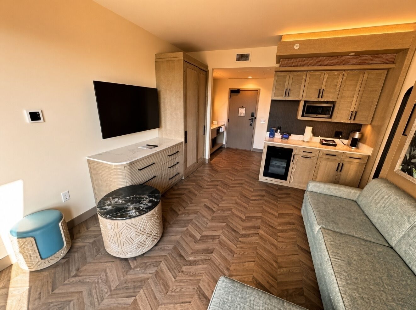A compact living space with a sofa, a television, a kitchen area, and a herringbone-patterned floor.