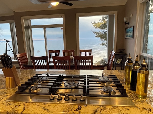 A kitchen with a gas stove, wooden chairs, and a view of the sea.