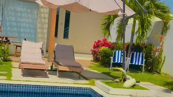 Piscina com espreguiçadeiras e um guarda-sol, cercada por um jardim bem cuidado e um edifício com varanda.
