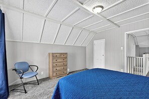 A bedroom with a sloped ceiling, a bed with a blue quilt, a wooden dresser, a chair, and a white door.