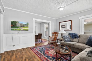 A living room with a wooden floor, a sofa, a coffee table, a rocking chair, a dining area with a table and chairs, a television mounted on the wall, and a window with blinds.