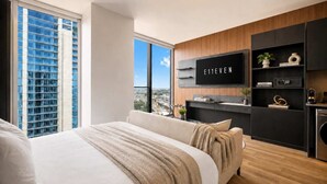 Ein modernes Schlafzimmer mit einem großen Bett, einem an der Wand befestigten Fernseher und Blick auf eine Stadtlandschaft durch ein großes Fenster.