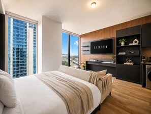 A modern bedroom with a large bed, a television mounted on the wall, and a view of a cityscape through a large window. - Elegant E11EVEN Residence with ELITE Amenities (Miami)