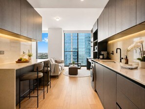 A modern kitchen with a central island, bar stools, and a view of a cityscape through large windows. - Elegant E11EVEN Residence with ELITE Amenities (Miami)
