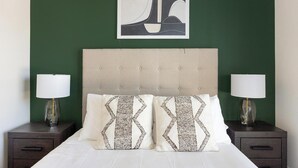 A neatly made bed with two decorative pillows, flanked by two bedside tables with lamps, against a green wall with a framed artwork above. - Landing | Modern 2BD, Game Room, Gym (Goodyear)