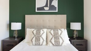 A neatly made bed with two decorative pillows, flanked by two bedside tables with lamps, against a green wall with a framed artwork above. - Landing | Modern 2BD, Game Room, Gym (Goodyear)