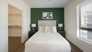 Ein Schlafzimmer mit einem Bett, zwei Nachttischen mit Lampen, einer grünen Akzentwand, einem gerahmten Kunstwerk über dem Bett und einem Fenster mit Jalousien.