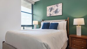 A neatly made bed with white linens, two blue pillows, and a headboard against a green wall. There are two bedside tables with lamps, and a framed artwork above the bed. - Landing | Incredible 1BD, Courtyard (Orlando)