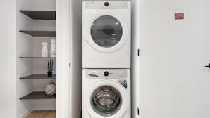 A compact laundry room with a stacked washer and dryer, shelves with toiletries, and a potted plant. - Landing | Incredible 1BD, Courtyard (Orlando)