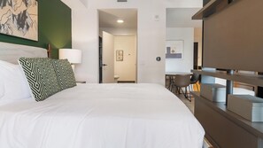 A hotel room with a bed, a pillow, a nightstand, a lamp, a painting on the wall, and a view into the dining area. - Landing | Stunning Studio, Pool, Gym (Orlando)