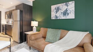 A modern living room with a sofa, a lamp, and a kitchen area with appliances. - Landing | Stunning Studio, Pool, Gym (Orlando)