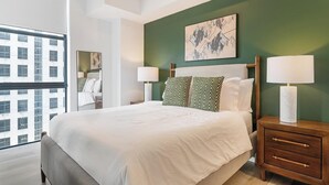 Una habitación de hotel con una cama grande, dos mesitas de noche con lámparas, una pared de acento verde y una ventana con vistas a edificios.