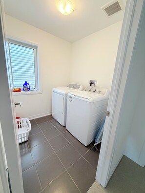 Une buanderie équipée de deux machines à laver et de deux sèche-linge à chargement frontal, tous deux de couleur blanche, d’une fenêtre avec des stores, d’un panier à linge et d’une bouche d’aération au plafond.