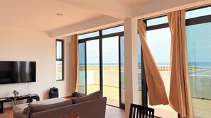 A modern living room with a flat-screen TV, a sofa, a table with a speaker, and large windows with curtains. - Joli Penthouse 60m² Rooftop Terrace, 360° View (Sal)