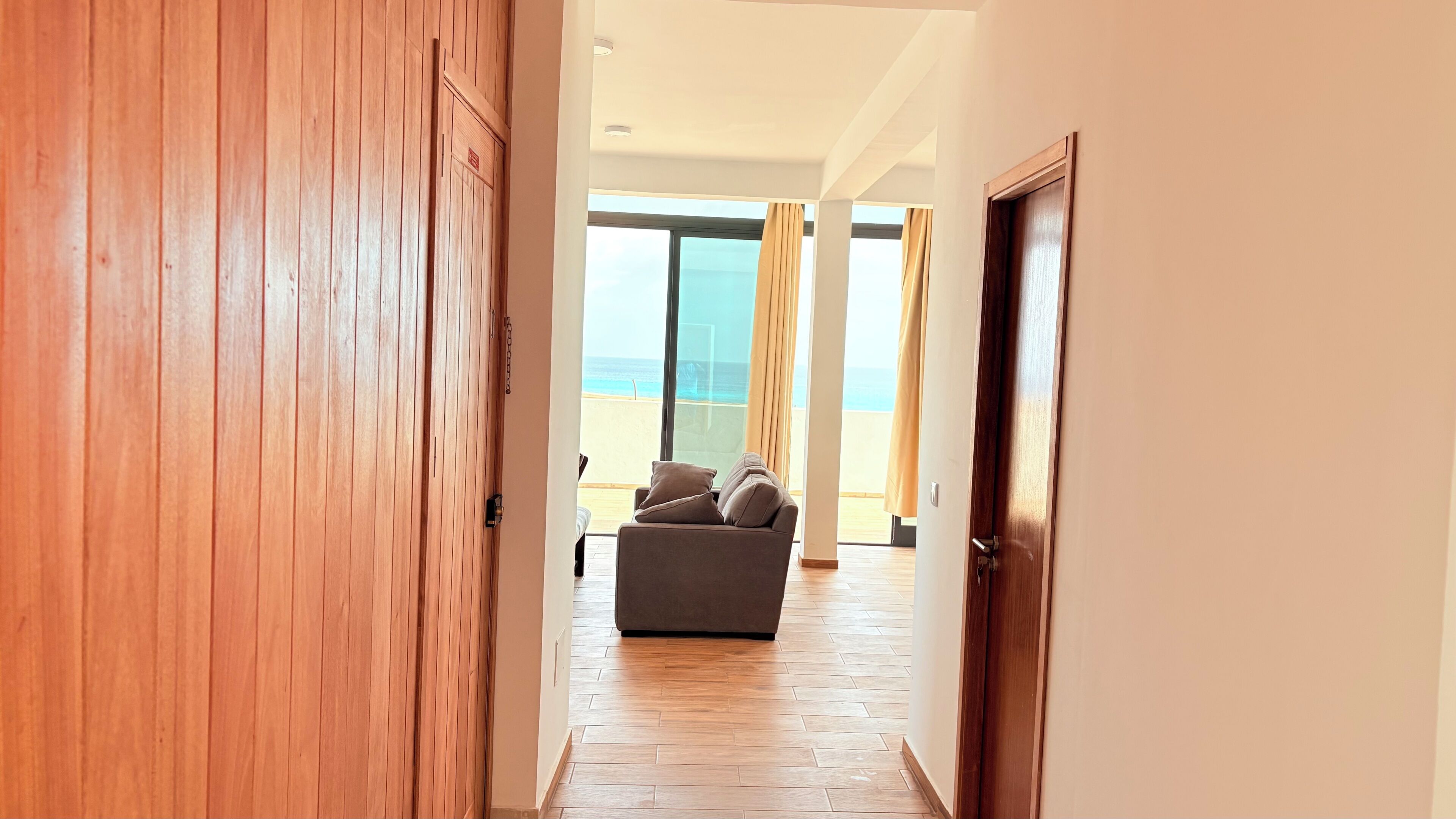 A hallway leading to a room with a view of the beach, wooden flooring, and a grey sofa.