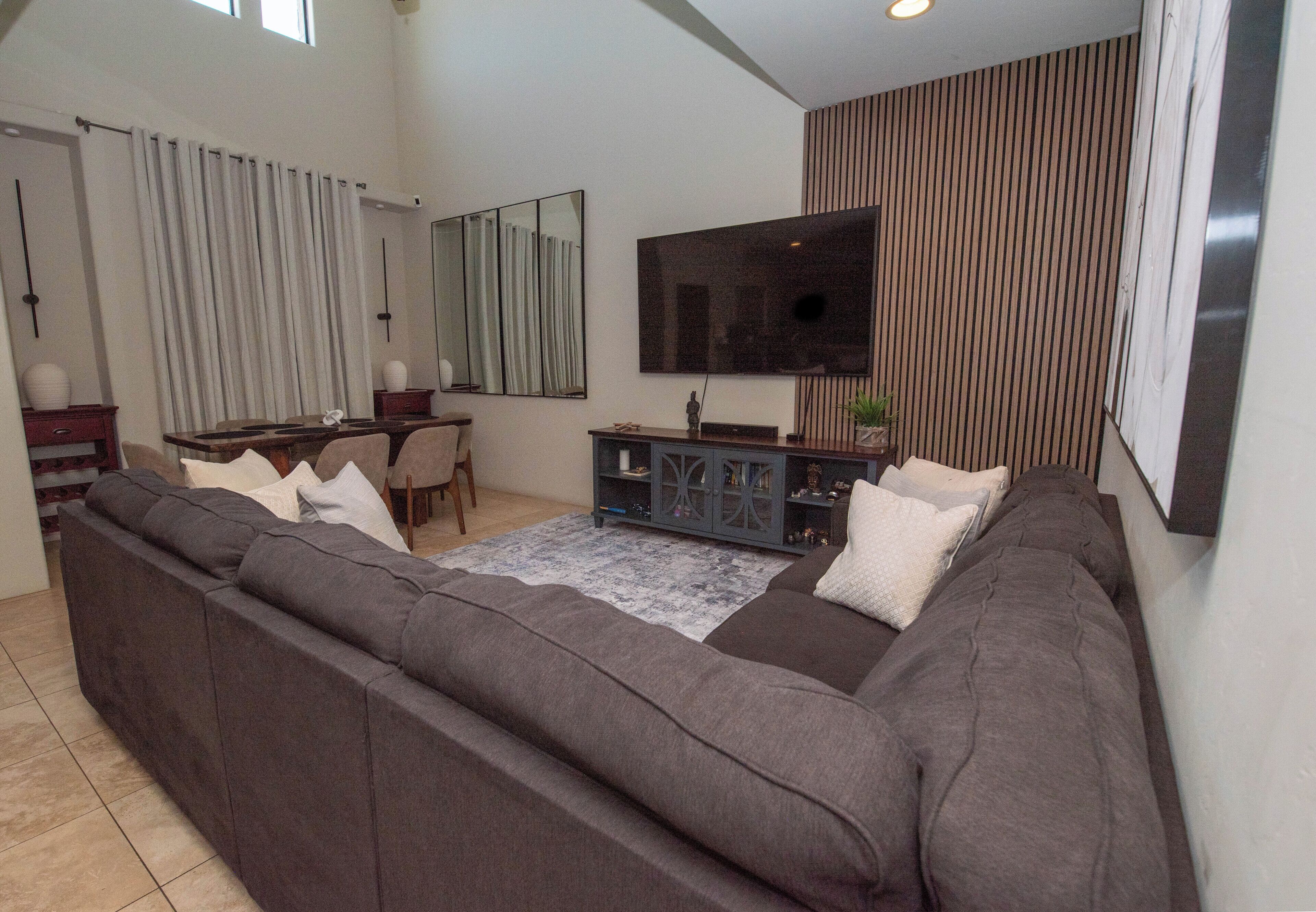 A modern living room with a large gray sectional sofa, a flat-screen TV mounted on the wall, and a dining area with a wooden table and chairs.