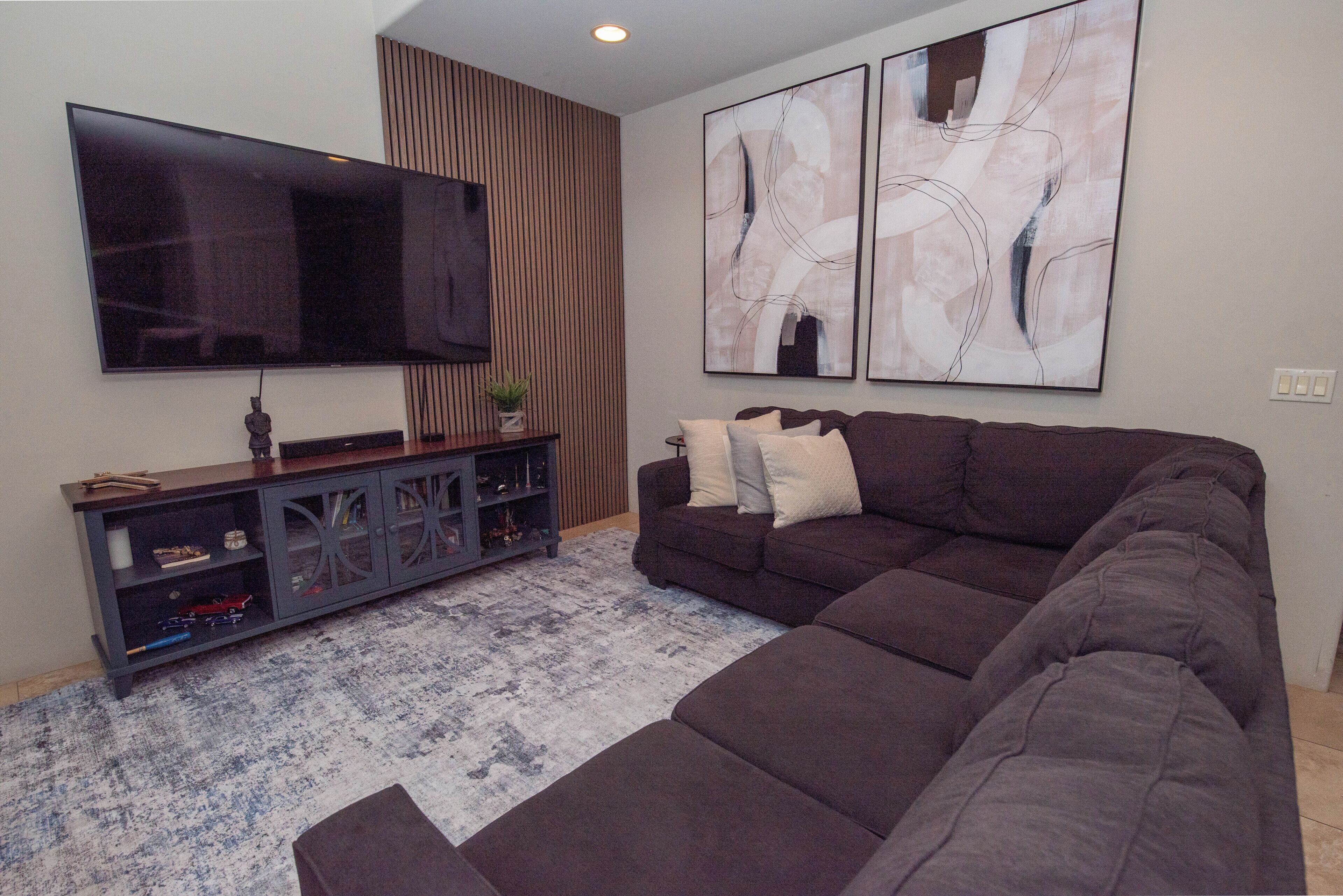 Un salon comprenant un canapé d’angle, un téléviseur à écran plat fixé au mur, un meuble décoratif avec des portes en verre et des œuvres d’art abstraites accrochées au mur.