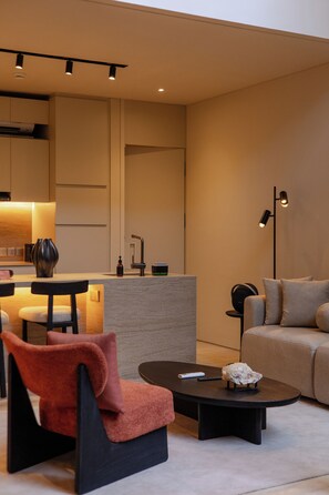 Una sala moderna con un sofá, una mesa de centro y un sillón. Se observa una cocina al fondo, con un fregadero y una encimera.