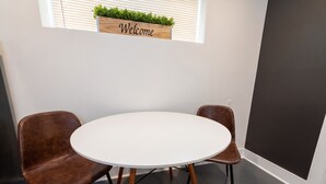 Une salle de réunion avec une table ronde et deux chaises marron, une pancarte « Bienvenue » au-dessus de la porte, et une fenêtre avec des stores.