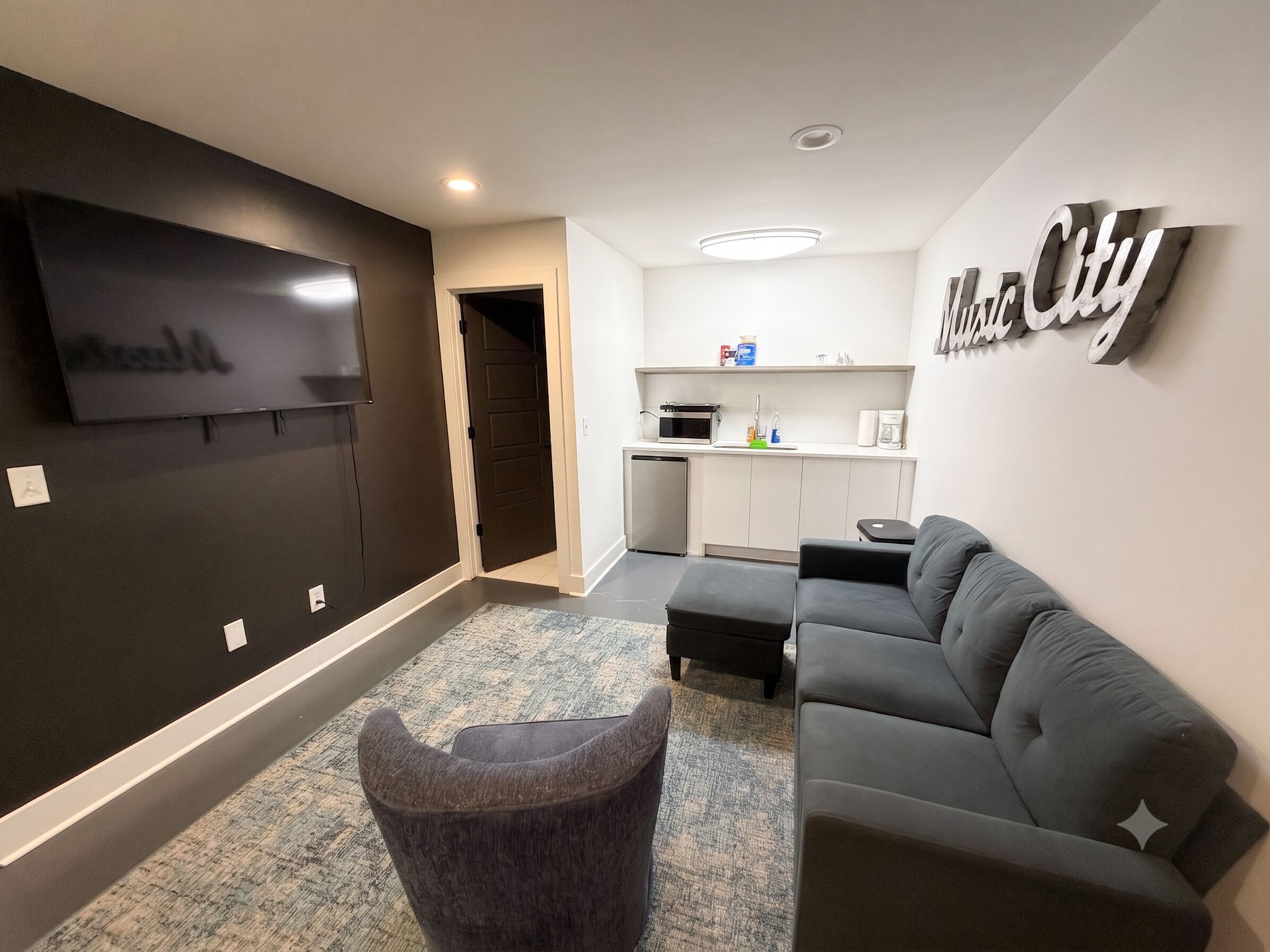 A modern living room with a flat-screen TV, a grey sofa, a small ottoman, and a kitchen area with a microwave and coffee maker.