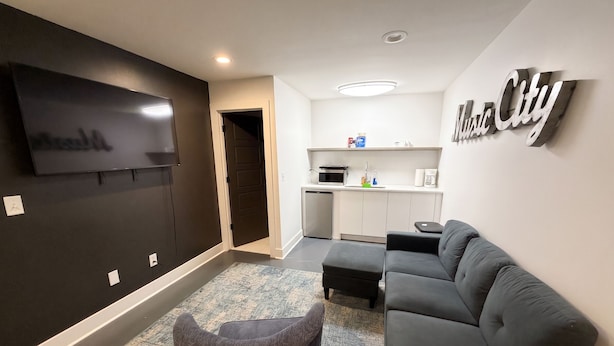 A modern living room with a flat-screen TV, a grey sofa, a small ottoman, and a kitchen area with a microwave and coffee maker.