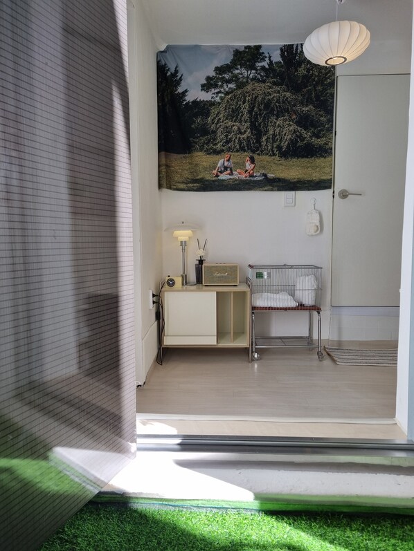 A room with a bed, a bedside table with a lamp, a radiator, a window with blinds, and a door.