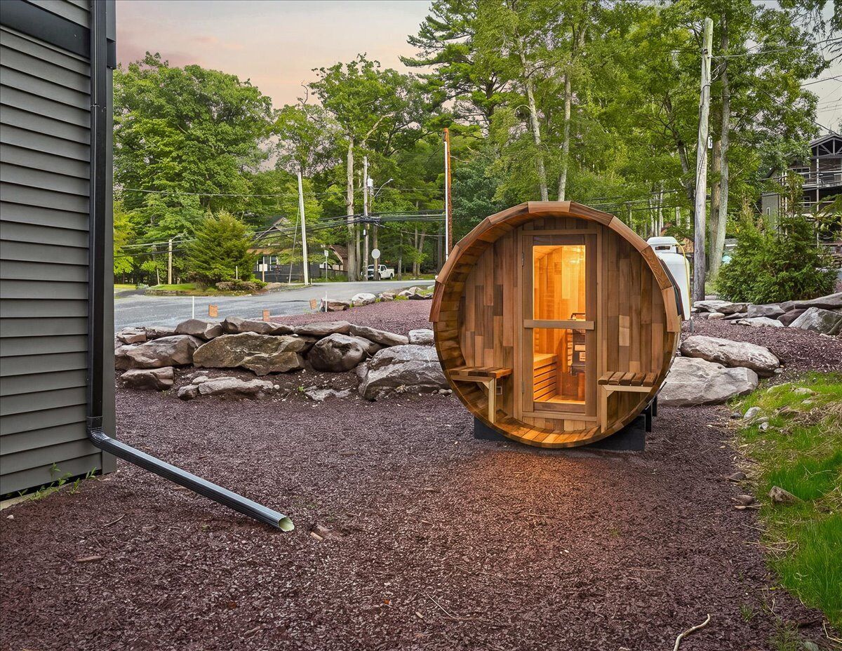 A wooden barrel structure with a door, positioned on a gravel surface with rocks and trees in the background.