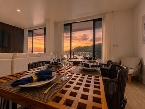 Un coin repas avec une table dressée pour deux personnes, de grandes fenêtres offrant une vue sur le coucher du soleil et un canapé en arrière-plan.