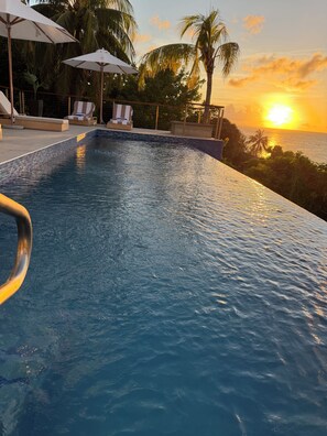 Una piscina con vista al atardecer sobre el océano, sillas de descanso y sombrillas.