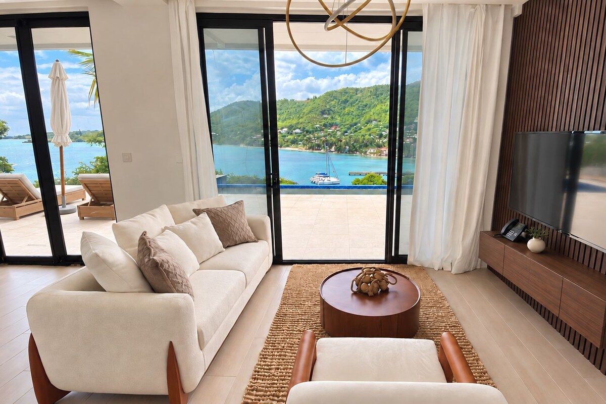 Una sala de estar moderna con un sofá, una mesa de centro y un televisor. Hay un ventanal con vistas a una playa y al mar.