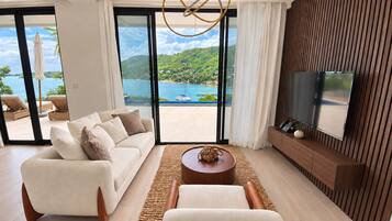 A modern living room with a sofa, coffee table, and a TV. There is a large window with a view of a beach and the sea.