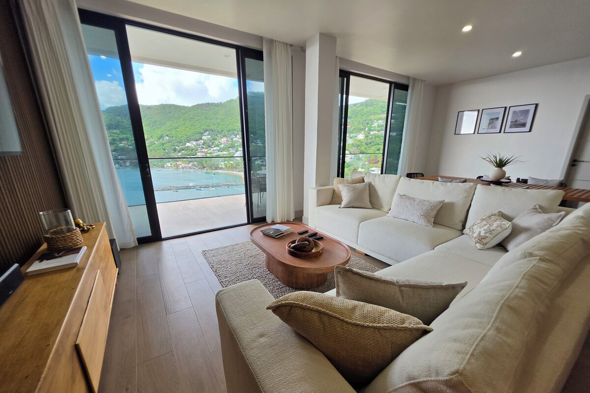 Una sala de estar moderna con un sofá, una mesa de centro y vista a la costa a través de ventanas amplias.