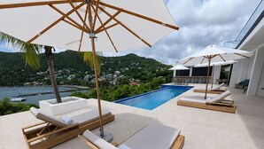 Zona junto a la piscina con tumbonas y sombrillas, con vista despejada al mar y colinas al fondo.