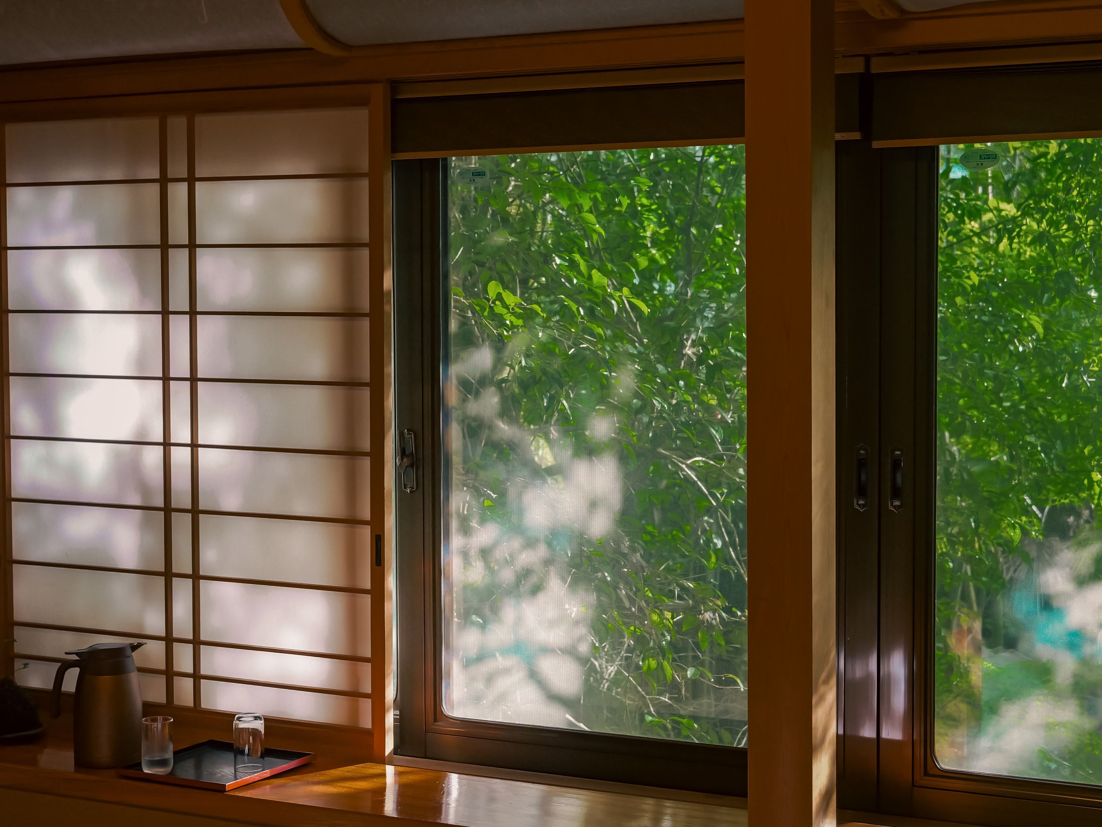 Um quarto japonês tradicional com tatames, um painel shoji e vista para o verde através de uma grande janela.